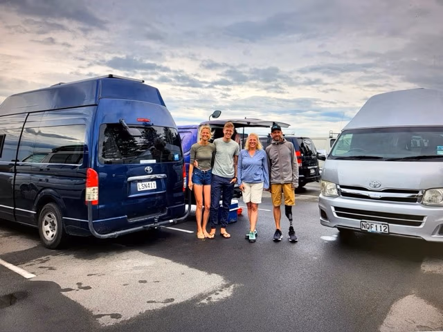 Four people stand smiling between two camper vans from BANZ Travelcars New Zealand Campervan Hire in a parking lot under a cloudy sky. The group appears relaxed and casual. The vans are blue and silver.