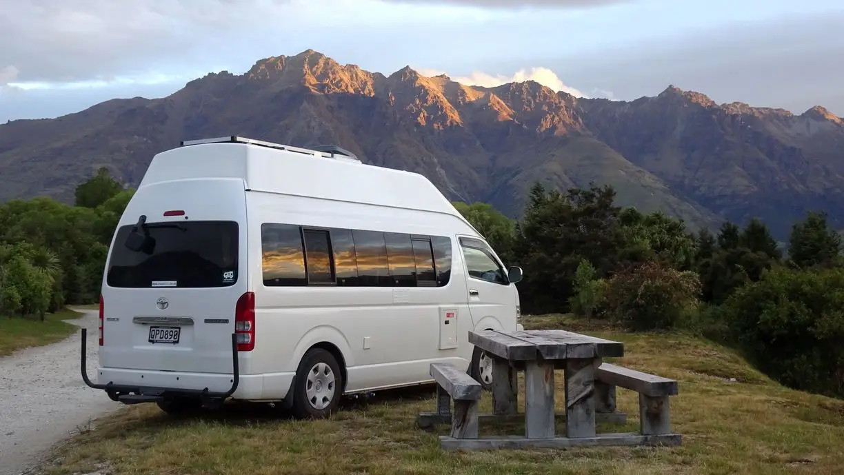 A BANZ camper van is parked beside a wooden picnic table on grassy terrain, with tall, sunlit mountains and green trees in the background. Hire a campervan in New Zealand to experience breathtaking views like this under a partly cloudy sky.