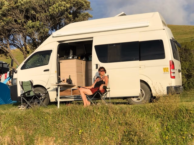 A woman relaxes in a camping chair outside her white BANZ camper van, enjoying a drink on a sunny day. Experience the freedom of the outdoors with self contained campervan New Zealand rental for your next adventure.
