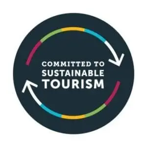 A circular logo with two white arrows forming a loop around the words Committed to Sustainable Tourism on a dark background, with segments of color along the circle—perfect for those seeking sustainable New Zealand campervan rentals.